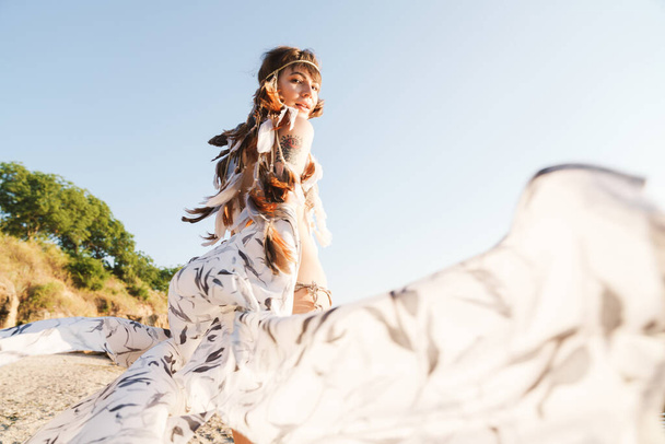 Image of hippie girl wearing feather headband walking by seaside - Fotó, kép