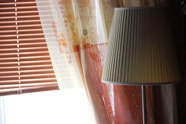 коричневые закрытые жалюзи, шторы и напольная лампа с бежевым оттенком. элементы интерьера дома в ясный солнечный день
 - Фото, изображение