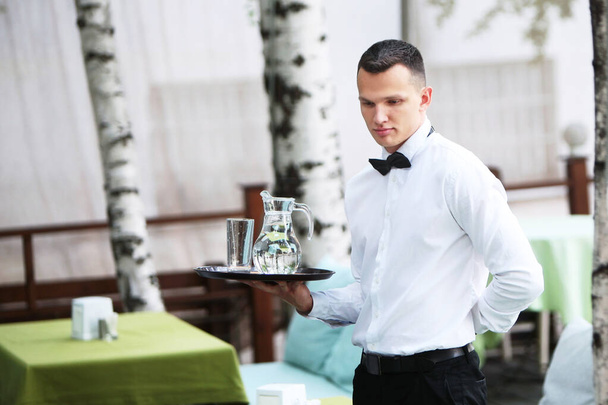 Ένας νεαρός, όμορφος σερβιτόρος κρατάει ένα δίσκο σε μια καράφα με νερό. Η έννοια της επιχείρησης εστιατόριο. Το προσωπικό στον τομέα των επιχειρήσεων εστιατορίων. - Φωτογραφία, εικόνα
