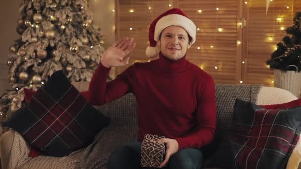 Portrait of Smiling Young Handsome Man Wearing Santas Hat and Winter Sweater Sitting on the Sofa Holding Present and Waving Hi with Christmas Tree at the Background. Concept of Holidays and New Year. - Video