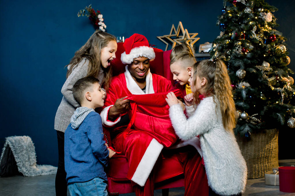 Five children look into the bag of African Santa Claus and laughing, African Santa Claus is seated and gives presents to beautiful little children in a christmas-decorated room. - Foto, immagini