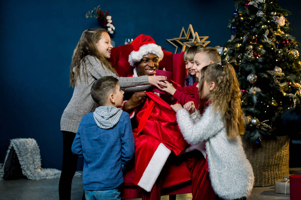 Five children look into the bag of African Santa Claus and laughing, African Santa Claus is seated and gives presents to beautiful little children in a christmas-decorated room. - Фото, зображення