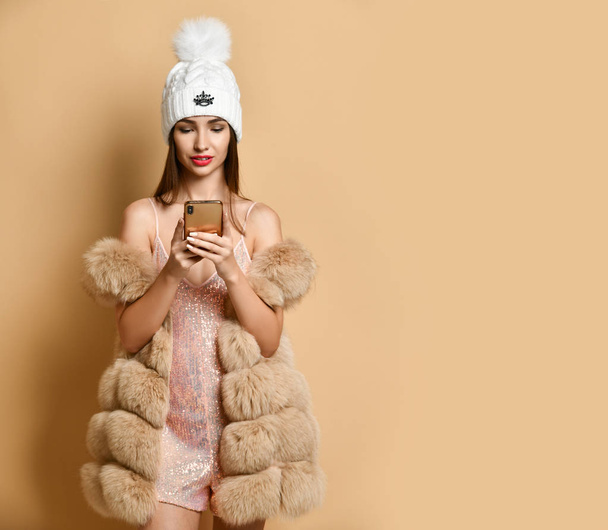 Young girl in party dress, fur vest is spending time with her smartphone, social networks, online shopping on beige - Zdjęcie, obraz