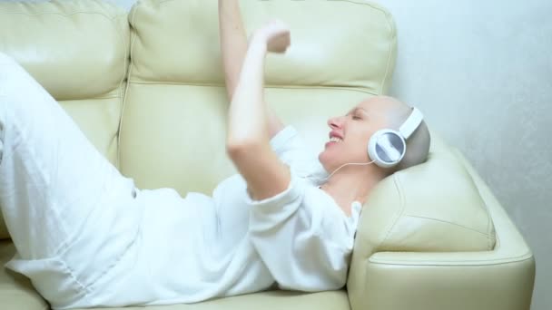 beautiful bald girl in headphones enjoys music at home on the couch - Filmmaterial, Video