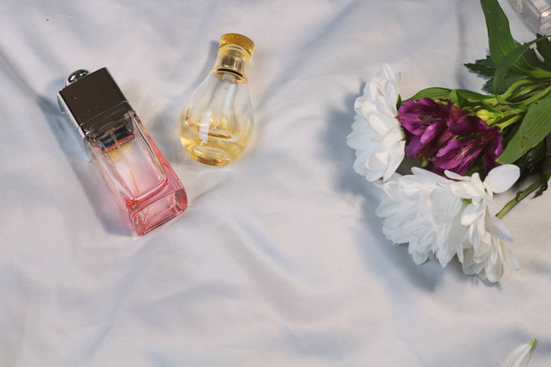 Many colorful fashion trendy bottles of perfume toilet water fragrances  lie on a silver gray sheet on bed next to cute gentle pink rose chamomile  flower, top view flat lay. Female, male fragrance - Photo, Image