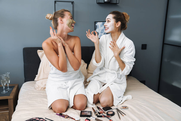 Cheerful young women taking care of their skin with face mask sit on bed. - Фото, изображение