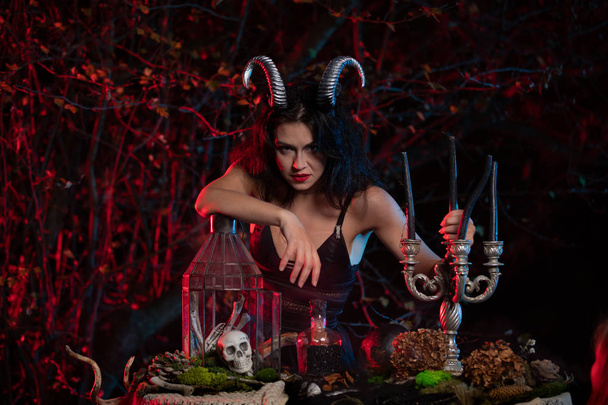 Gyönyörű boszorkány szarvakkal a fején készen áll, hogy boszorkányságot kövessen el az erdő hátterében. Halloween kép. - Fotó, kép