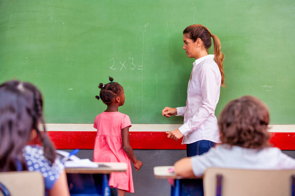 Afrykańska dziewczyna w podstawówce z nauczycielem przed zieloną tablicą. - Zdjęcie, obraz