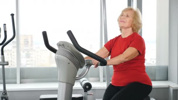 Portrét aktivní staré ženy ve fitness klubu zabývající se kardio tréninkem na cvičebním kole. Zdravá a aktivní stará žena. Aktivní maturanti. Stará paní ve fitku. Shot in 4k - Záběry, video