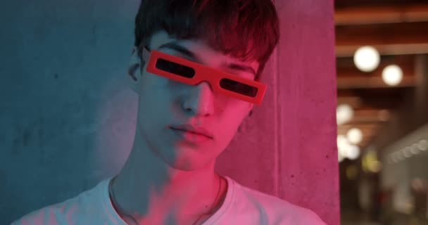 Portrait of Young Millenial Guy in Funny 3d Glasses Standing Leaned to Wall Turning his Head and Looking Straight to Camera at Futuristic Neon Club Lights. Close Up shot. - Filmati, video