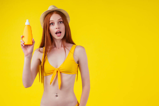 redhaired ginger woman in stylish bikini holding sunscreen bottle making the right choice in studio yellow background - Фото, изображение