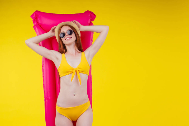 redhaired ginger female posing floating with inflatable mattress wearing swimmingsuit and hat,good looking in studio yellow background - Zdjęcie, obraz