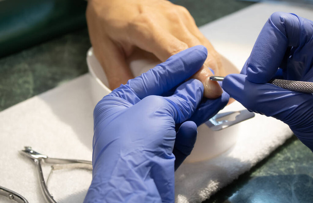 Male manicure. Woman hands in blue sterile gloves holding male finger and manicure instrument making manicure procedure. - Photo, image