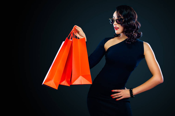 Black Friday sale concept for shop. Shopping woman in sunglasses holding red bag isolated on dark background. - 写真・画像