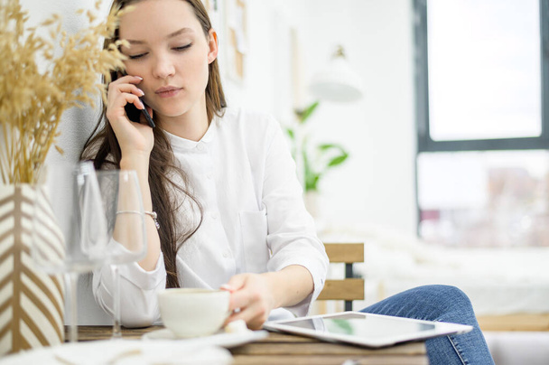 A woman in a white shirt is drinking coffee in a cafe. A female office worker at a lunch break solves a business problem by phone. Mid-level manager - Foto, afbeelding
