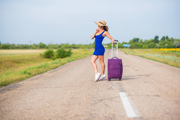 A woman is standing on the road with a hat and with a big bag. Girl in a blue tight-fitting dress with a purple suitcase on the roadway. A brunette with a sports figure is waiting for a bus. - Φωτογραφία, εικόνα