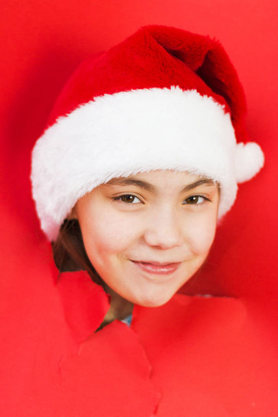 View of a girl in a Santa hat through a hole in the red paper. Smiling girl makes a hole in the paper. Festive Christmas background - Foto, imagen