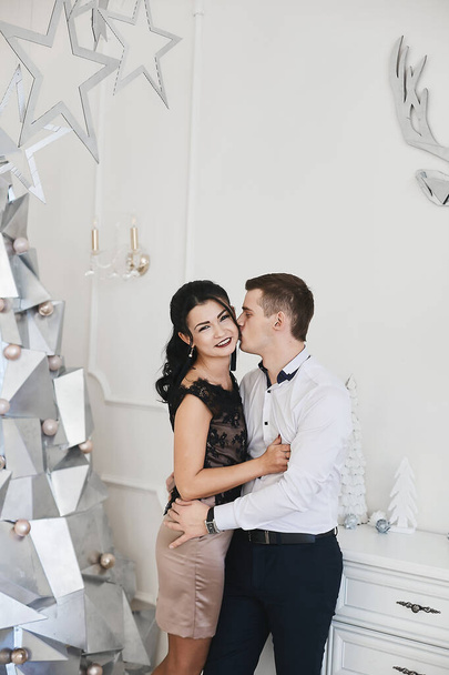 Beautiful couple in love hugging and kissing in Christmas interior. Young brunette woman in skirt and blouse hugging with handsome businessman in a white shirt. Loving married couple. New year - Photo, Image