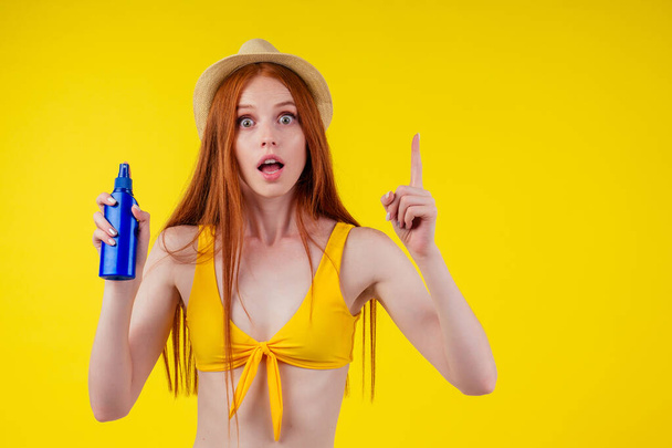 redhaired ginger woman in stylish bikini holding sunscreen bottle making the right choice in studio yellow background - Foto, Imagem