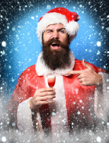handsome bearded santa claus man with long beard on smiling face holding glass of alcoholic shot in red christmas or xmas sweater and new year hat on blue studio backgrou - Фото, зображення