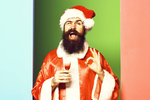 handsome bearded santa claus man with long beard on funny face holding glass of alcoholic shot in red christmas or xmas coat and new year hat on colorful studio backgrou - Foto, immagini
