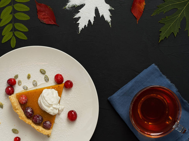 Slice of homemade traditional American pumpkin pie decorated with cream and cranberries on a white plate. Top view. On a black background multi-colored autumn leaves and a cup with tea. - Photo, Image
