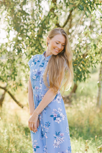 Een blonde in een blauwe zomerjurk met lang haar glimlacht en kijkt naar beneden - Foto, afbeelding