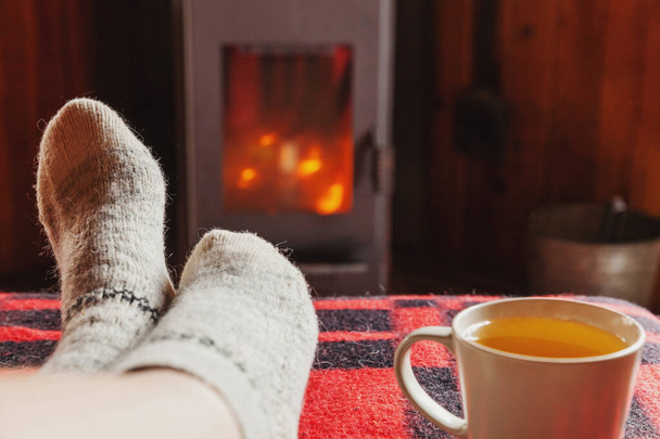Nohy v zimních šatech s vlnohami a šálkem čaje u krbu doma v zimě nebo v podzimní noci relaxující a zahřívací - Fotografie, Obrázek