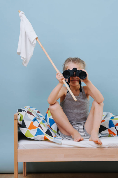 Little boy is sitting on unmade bed with white flag, looking through binoculars - Photo, image