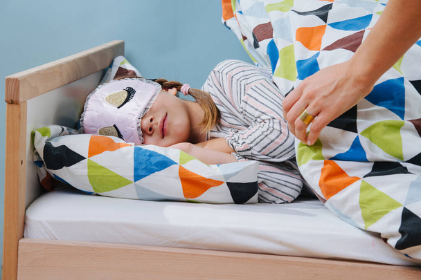 Mother tucks her little daughter in bed. Girl wearing sleeping mask. - Foto, Imagen