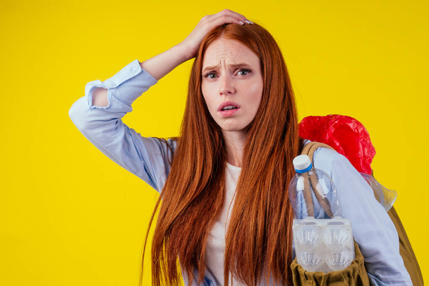 redhaired ginger european woman with cotton eco bag full of plastic trash in yellow studio background.recycling ecology solution concept - Zdjęcie, obraz