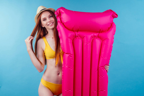redhaired ginger female good-looking wearing stylish yellow swimmingsuit holding inflatable pink mattress in studio blue background - Photo, Image