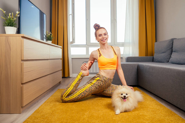 redhaired ginger woman doing yoga asana at day light living room on the yellow carpet with her cute white fluffy spitz - Photo, image
