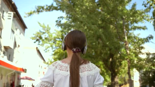 girl with a backpack goes to park in headphones and listens to music and smiles, teenager happily waves his hand at camera lens. come after me, girl travels around city. Slow motion. - Séquence, vidéo
