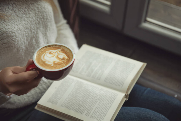 Woman with cup of coffee reading book near window indoors, closeup - Foto, imagen