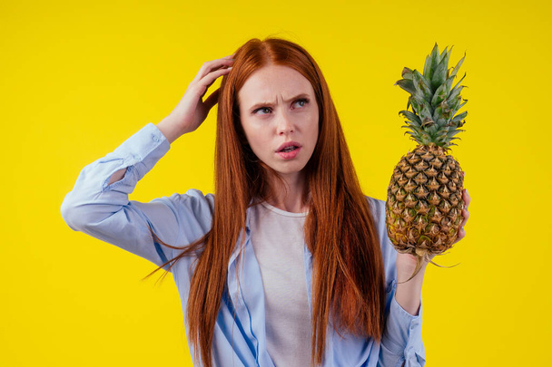 redhaired ginger surprised woman choosing pineapple in studio yellow background - Fotó, kép