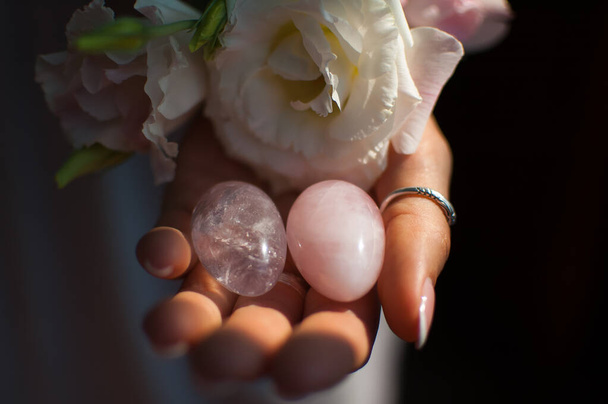 Женские руки, держащие в руках два яйца йони для вамфита, имбилдинга или медитации, сделаны из розового кварца и прозрачного фиолетового аметист с белыми цветами в помещении
 - Фото, изображение