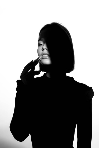 Divatreklám portré egy fiatal, gyönyörű lányról, aki rúzst használ. Fekete-fehér sziluett epikus fénnyel. - Fotó, kép