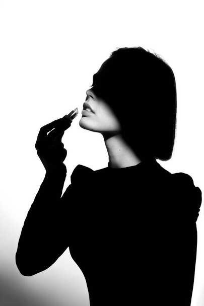 Fashion advertising portrait of a young beautiful girl using lipstick. Black and white silhouette with epic light. - Photo, Image