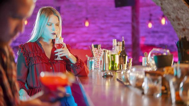 Linda jovem loira sentada ao lado do barman stand - bebendo uma bebida da palha - um homem sentado em seu telefone em primeiro plano
 - Foto, Imagem