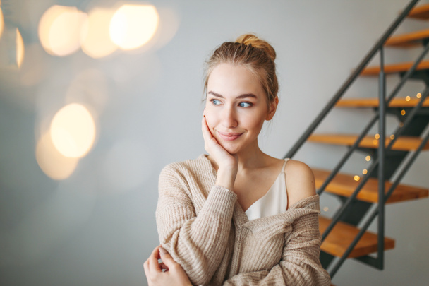 Young beautiful thoughtful girl with blonde hair in an silk dress and cozy cardigan dreaming about something at home against background of bokeh - Photo, Image