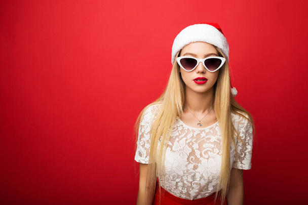 Pensive blonde in santa hat on a red isolated background. White rimmed sunglasses. - Foto, afbeelding