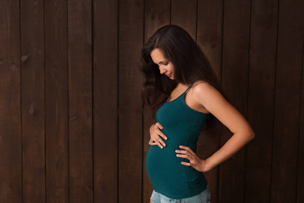Pregnant woman in jeans shorts and green top holds hands on belly on a dark brown background. Pregnancy, maternity, preparation and expectation concept. Beautiful tender mood photo of pregnancy - Fotografie, Obrázek