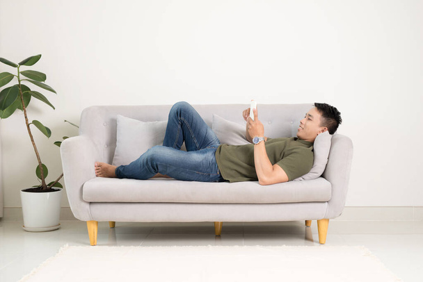 Uomo rilassato utilizzando uno smartphone sdraiato su un divano / divano in soggiorno a casa
 - Foto, immagini
