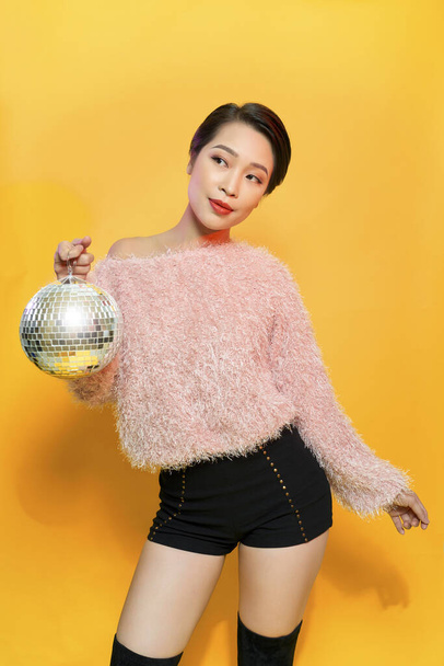 Portrait of charming young female showing tongue and fooling around holding mirror ball on head posing on yellow background. Party and glitter fashion concept - Photo, Image