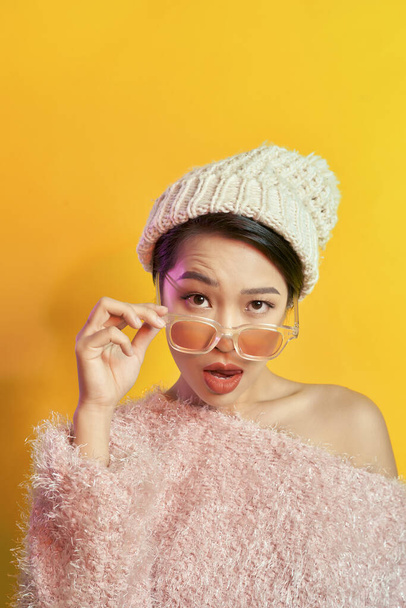 Ragazza asiatica emotiva con trucco colorato in posa su sfondo giallo con occhiali. Foto indoor di donna stupita in pelliccia alla moda
. - Foto, immagini