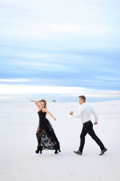 Joven mujer rubia y hombre corriendo en la estepa de invierno, vistiendo vestido negro
. - Foto, imagen