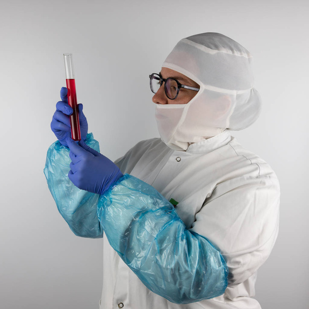 Young woman with glasses, wearing white, sterile, protective clothing and blue gloves, holding a tube with a sample, red liquid for food industry, pharmacy, Quality control concept - Photo, image