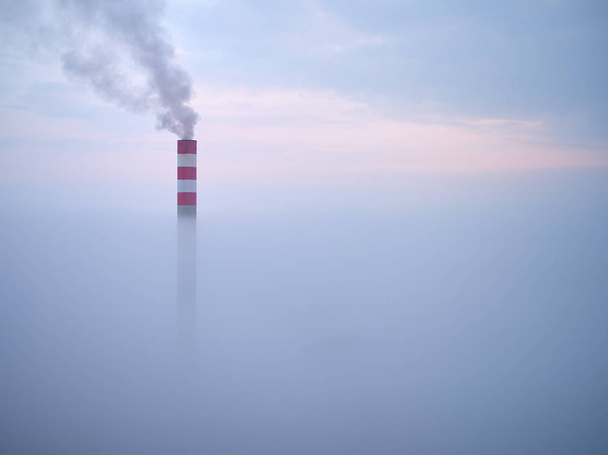 Smoking chimney of a power plant coming out of the inverse fog. The power plant chimney in operation, a red-white striped, rises above the surrounding fog. Energy industry. Heating plant in operation. - 写真・画像