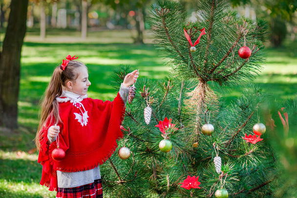 Pienen tytön muotokuva joulukuusen lähellä. Tyttö koristaa joulukuusi metsässä joulupallot
. - Valokuva, kuva
